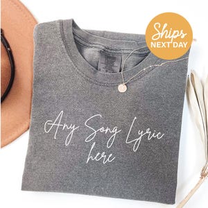 A gray t-shirt with the text "Any Song Lyric here" printed on it, along with a necklace and a hat.