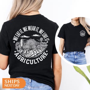 May include: Black t-shirt with a circular graphic featuring a farm scene with a barn, windmill, and fields. The text around the graphic reads "WE EAT IT, WE WEAR IT, WE LIVE IT AGRICULTURE". The shirt is worn by a person.