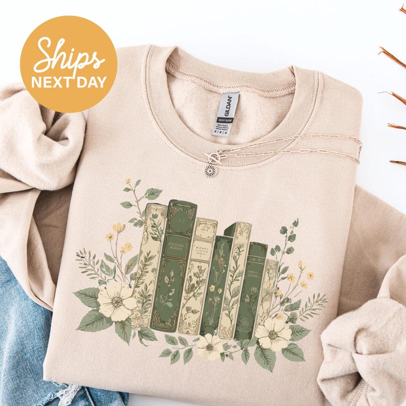 A beige sweater with a floral design of books and flowers, along with a necklace and a circular orange sign that reads "Shops NEXT DAY".