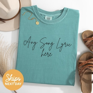 A turquoise t-shirt with the text "Any Song Lyrics here" printed on it, along with a beige hat, a gold necklace, and a pair of brown sandals, all laid out on a white surface.