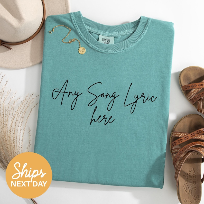 A turquoise t-shirt with the text "Any Song Lyrics here" printed on it, along with a beige hat, a gold necklace, and a pair of brown sandals, all laid out on a white surface.