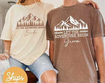 Puede incluir: Dos camisetas a juego, una beige y otra marrón, con un diseño de montaña y bosque y el texto "LET THE ADVENTURE BEGIN". La camiseta beige dice "Bride", y la camiseta marrón dice "Groom".