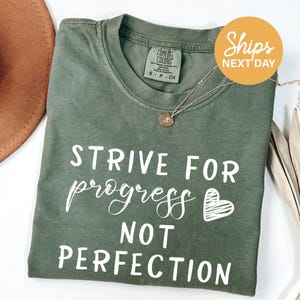 a green t-shirt with white text that reads "STRIVE FOR PROGRESS NOT PERFECTION". The t-shirt is folded and placed on a white surface, with a brown hat and a necklace visible in the background.