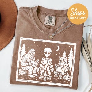 May include: Brown Comfort Colors t-shirt featuring a white graphic of Bigfoot, an alien, and an owl reading books around a campfire. The shirt has a crew neck and short sleeves. A silver necklace with a pendant is also visible.