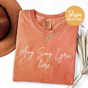 a pink t-shirt with the text "Any Song Lyric here" written on it, along with a necklace and a hat.