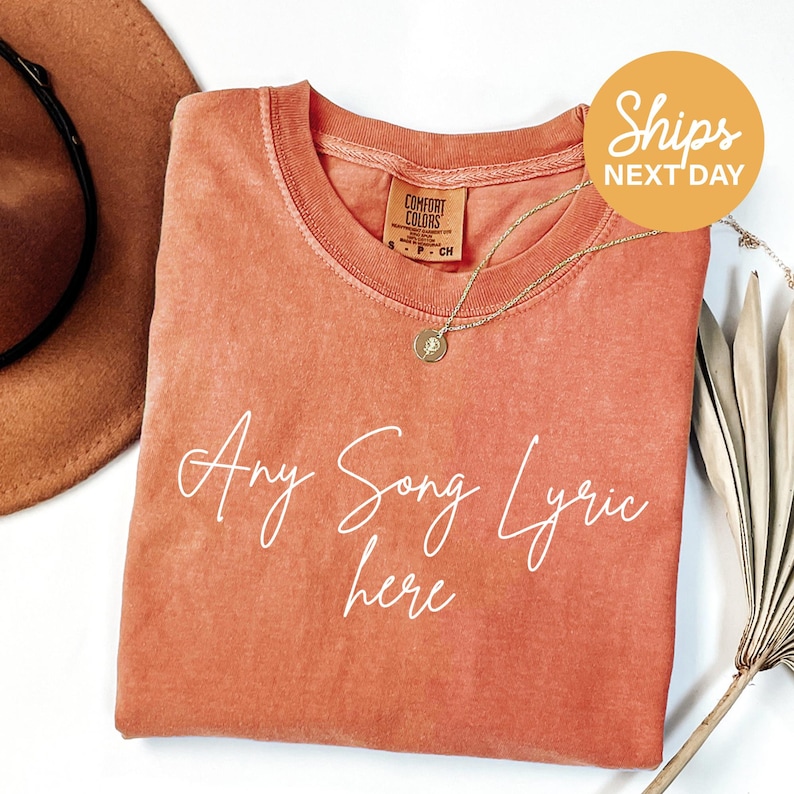 a pink t-shirt with the text "Any Song Lyric here" written on it, along with a necklace and a hat.