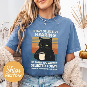 A woman wearing a blue t-shirt with a graphic of a black cat holding a coffee cup and text that reads "I HAVE SELECTIVE HEARING I'M SORRY YOU WEREN'T SELECTED TODAY TOMORROW ISN'T LOOKING GOOD EITHER".