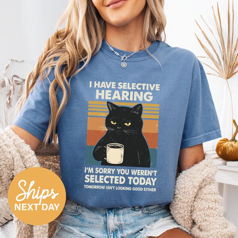 A woman wearing a blue t-shirt with a graphic of a black cat holding a coffee cup and text that reads "I HAVE SELECTIVE HEARING I'M SORRY YOU WEREN'T SELECTED TODAY TOMORROW ISN'T LOOKING GOOD EITHER".