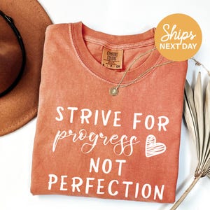 a red t-shirt with white text that says "STRIVE FOR PROGRESS NOT PERFECTION". The t-shirt is laid out on a white surface, with a brown hat and a plant visible in the background.