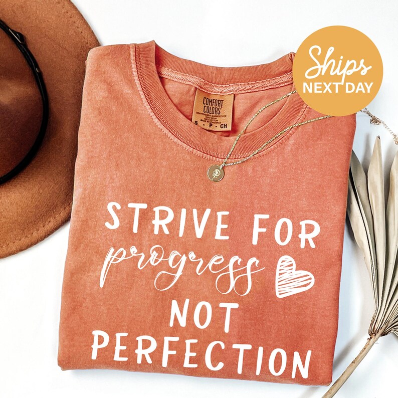 a red t-shirt with white text that says "STRIVE FOR PROGRESS NOT PERFECTION". The t-shirt is laid out on a white surface, with a brown hat and a plant visible in the background.