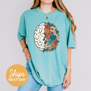 A woman wearing a turquoise t-shirt with a graphic design of a brain surrounded by colorful flowers.