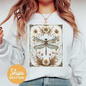 May include: A light gray sweatshirt featuring a detailed dragonfly design. The dragonfly is surrounded by floral elements in shades of beige, brown, and yellow. The design is framed with a decorative border. The text "Ships Next Day" is in a yellow circle.