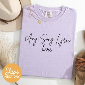 a purple t-shirt with the text "Any Song Lyric here" printed on it, along with a beige hat and a pair of sandals.