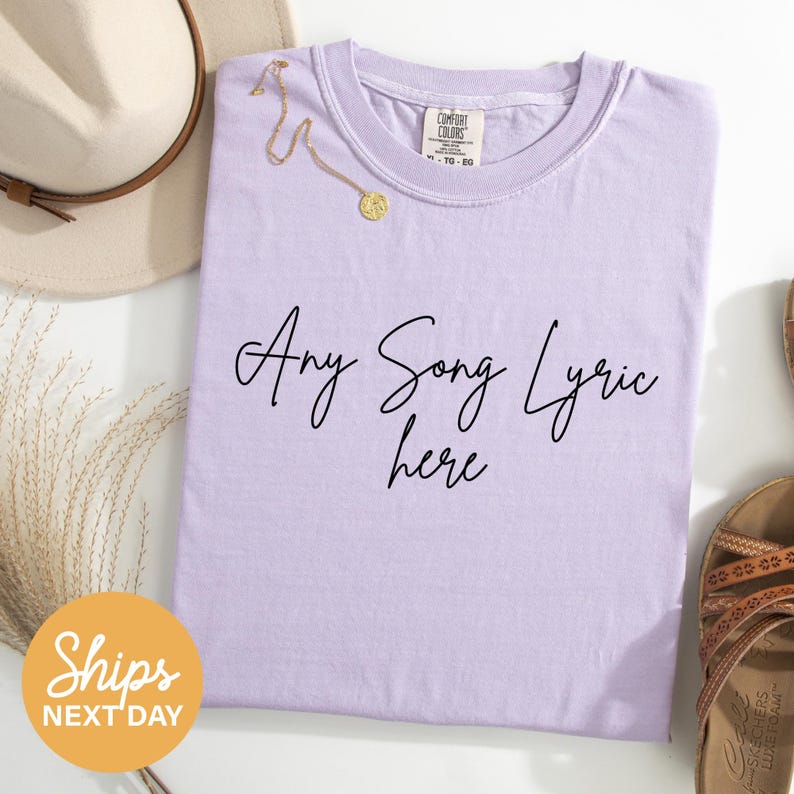 a purple t-shirt with the text "Any Song Lyric here" printed on it, along with a beige hat and a pair of sandals.