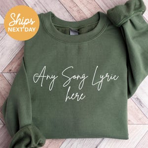 May include: An olive green sweatshirt with the text "Any Song Lyric here" in a white script font. The sweatshirt has a crew neck and long sleeves. A yellow circle with the text "Ships NEXT DAY" is in the upper left corner.