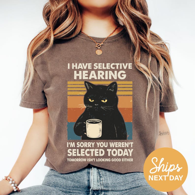 A woman wearing a t-shirt with a graphic design of a black cat holding a coffee mug and text that reads "I HAVE SELECTIVE HEARING I'M SORRY YOU WEREN'T SELECTED TODAY TOMORROW ISN'T LOOKING GOOD EITHER".
