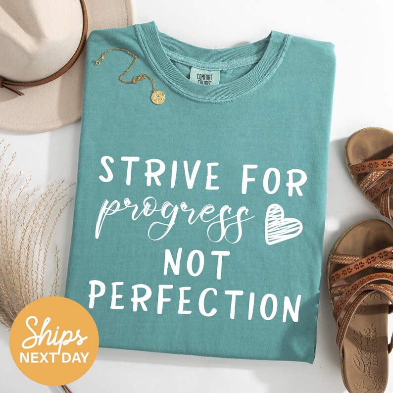 a turquoise t-shirt with the text "STRIVE FOR PROGRESS NOT PERFECTION" printed on it, along with a heart symbol. The t-shirt is laid out on a white surface, accompanied by a beige hat, a pair of brown sandals, and some dried plants.