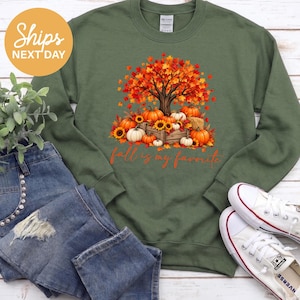 May include: An olive green crewneck sweatshirt featuring a graphic of a tree with vibrant orange and red autumn leaves, surrounded by pumpkins and sunflowers in a wooden crate, with the orange text fall is my favorite below.