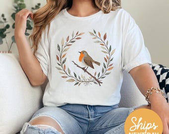 May include: White t-shirt featuring a watercolor illustration of a robin perched on a branch within a wreath of leaves. The bird has a red breast and brown feathers. The shirt is a relaxed fit and has short sleeves. The text "Ships Next Day" is in an orange circle.