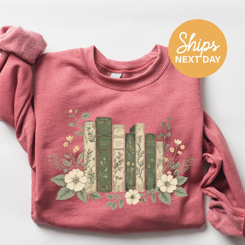 A pink sweatshirt with a graphic design of a row of books surrounded by flowers and leaves.