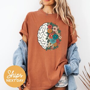 A woman wearing a brown t-shirt with a graphic of a brain surrounded by colorful flowers, standing against a white background.
