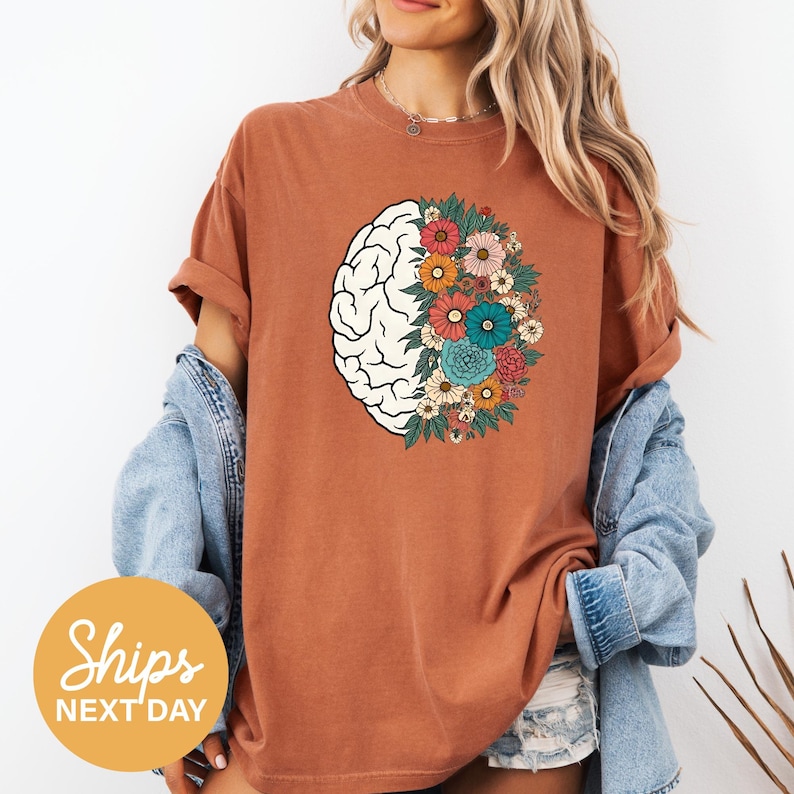 A woman wearing a brown t-shirt with a graphic of a brain surrounded by colorful flowers, standing against a white background.