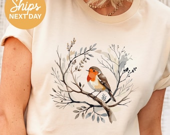 May include: Cream-colored t-shirt featuring a watercolor illustration of a robin perched on a branch within a wreath of twigs and leaves. The bird has a red breast and brown wings. The text "Ships Next Day" is in a yellow circle.