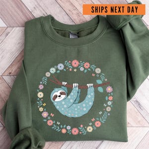 May include: A green sweatshirt with a floral wreath design featuring a blue sloth hanging from a branch. The sloth has a white and blue patterned design.