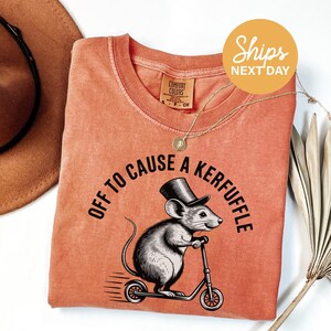 May include: Coral-colored t-shirt with a graphic of a mouse in a top hat riding a scooter. The text "OFF TO CAUSE A KERFUFFLE" arches above the mouse. A gold necklace and a brown hat are also visible.