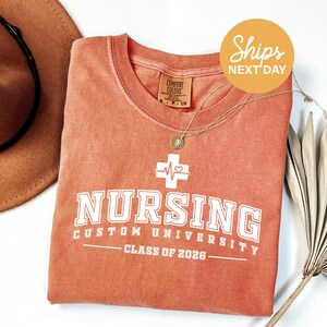 May include: A coral-colored t-shirt with the word "NURSING" in white block letters. Below the word "NURSING" is "CUSTOM UNIVERSITY" and "CLASS OF 2026". A gold necklace with a pendant is draped over the shirt. The shirt has a crew neck.