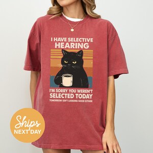 A red t-shirt with a graphic design depicting a black cat holding a coffee cup, accompanied by text that reads "I HAVE SELECTIVE HEARING I'M SORRY YOU WERE'N'T SELECTED TODAY TOMORROW ISN'T LOOKING GOOD EITHER".