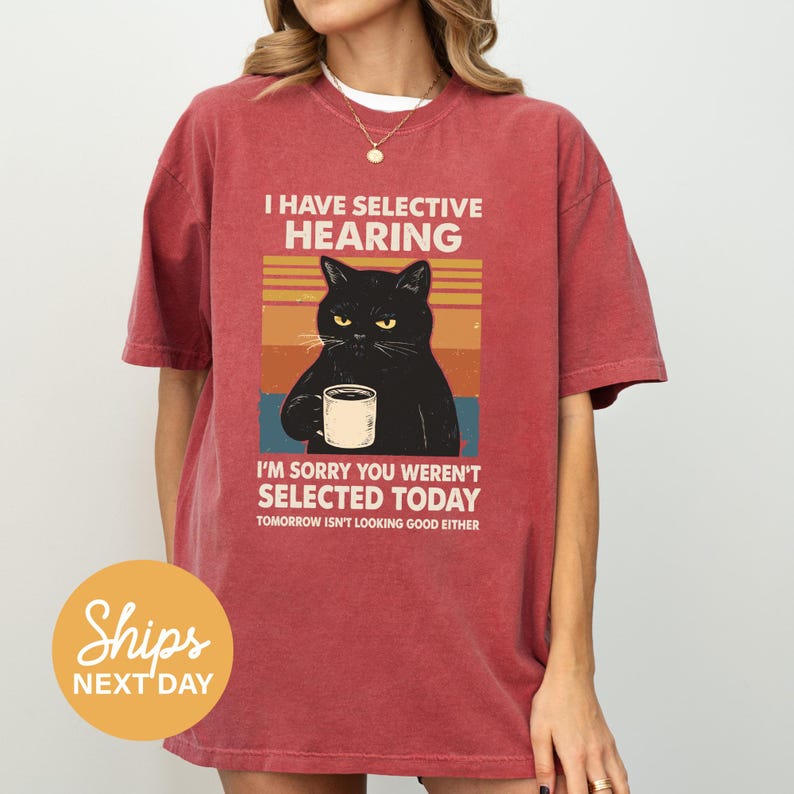 A red t-shirt with a graphic design depicting a black cat holding a coffee cup, accompanied by text that reads "I HAVE SELECTIVE HEARING I'M SORRY YOU WERE'N'T SELECTED TODAY TOMORROW ISN'T LOOKING GOOD EITHER".