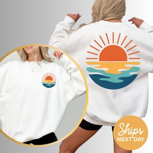 a woman wearing a white sweatshirt with a sunset on it