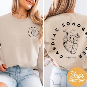 May include: Beige crewneck sweatshirt with a black graphic of a heart and the words "CARDIAC SONOGRAPHER" in a circular design. The front features a smaller version of the design. The sweatshirt is a neutral color.