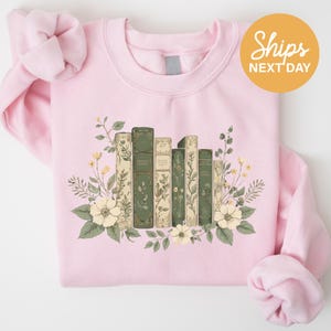 A pink sweatshirt with a graphic design of a row of books surrounded by flowers and leaves.