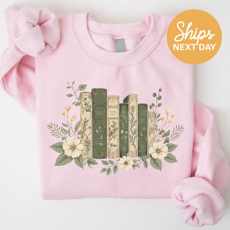 A pink sweatshirt with a graphic design of a row of books surrounded by flowers and leaves.