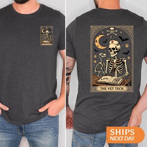 Peut inclure: T-shirt gris fonc&eacute; avec un motif de carte de tarot. L'avant pr&eacute;sente un squelette lisant sous un croissant de lune. Le dos montre un technicien v&eacute;t&eacute;rinaire squelette avec un st&eacute;thoscope, entour&eacute; de chauves-souris et d'&eacute;toiles. Le texte dit "THE VET TECH".