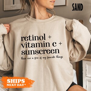 May include: A beige sweatshirt with black text that reads "retinol + vitamin c + sunscreen these are a few of my favorite things".