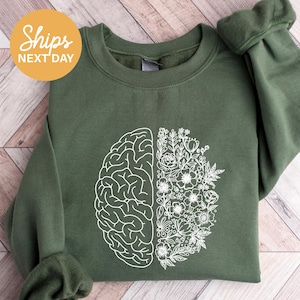May include: An olive green sweatshirt with a white graphic design. The design is split in half, with one side depicting a brain and the other side a floral arrangement. The text "Ships Next Day" is in a yellow circle in the upper left corner.