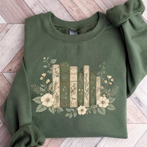 A green sweatshirt with a floral design featuring a row of books surrounded by flowers and leaves.