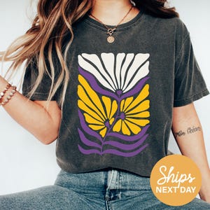 May include: Dark gray t-shirt featuring a floral design in white, yellow, and purple. The graphic has a retro aesthetic. The shirt is a crew neck and has short sleeves. The shirt is made of a soft material.