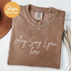 May include: A brown t-shirt with the text "Any Song Lyric here" in white script. The shirt has a crew neck and short sleeves. A silver necklace with a pendant is draped across the shirt. The shirt is a Comfort Colors brand.