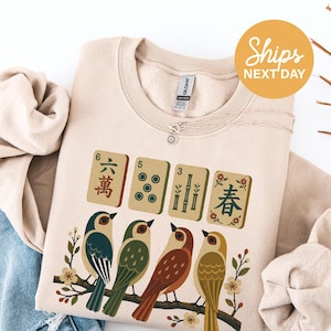 May include: A beige crewneck sweatshirt featuring a graphic print of four mahjong tiles above four colorful birds perched on a flowering branch. The tiles display the numbers 6, 5, 3, and a floral character. The sweatshirt has a ribbed collar and cuffs.