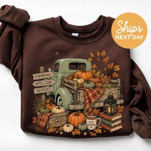 May include: A dark brown crewneck sweatshirt featuring a graphic illustration of a vintage green pickup truck loaded with pumpkins and autumn leaves. The design includes wooden signs labeled PUMPKINS, APPLE CIDER, and HAYRIDES, plus a lantern.