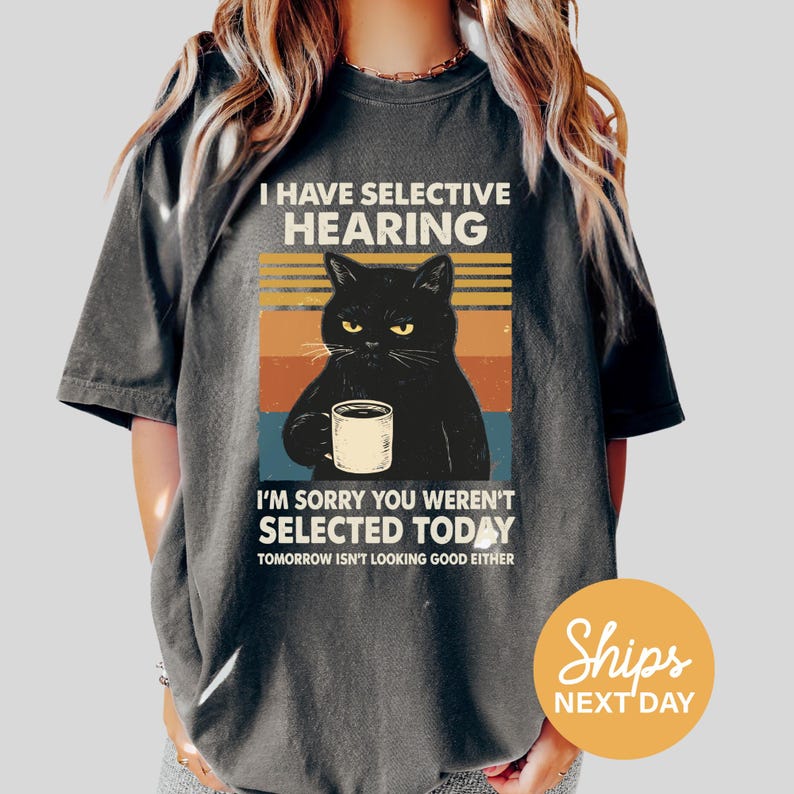 A person wearing a gray t-shirt with a graphic design depicting a black cat holding a cup and making a face, accompanied by text that reads "I HAVE SELECTIVE HEARING I'M SORRY YOU WEREN'T SELECTED TODAY TOMORROW ISN'T LOOKING GOOD EITHER".