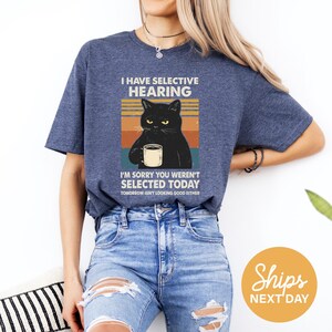 A woman wearing a blue t-shirt with a graphic of a black cat and text that says "I HAVE SELECTIVE HEARING" and "I'm sorry you weren't selected today, tomorrow isn't looking good either".