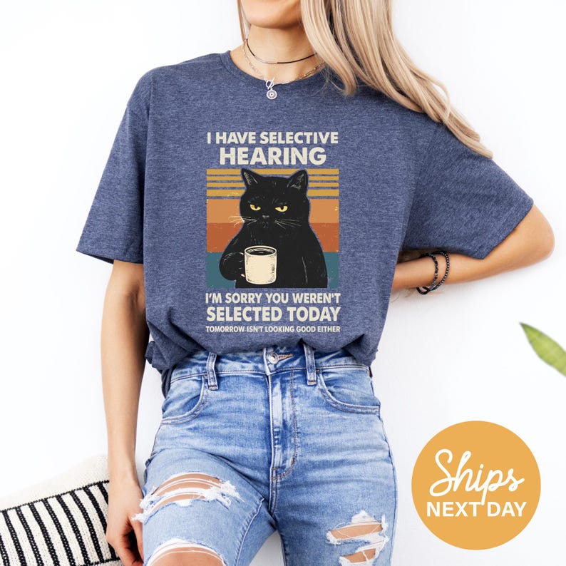 A woman wearing a blue t-shirt with a graphic of a black cat and text that says "I HAVE SELECTIVE HEARING" and "I'm sorry you weren't selected today, tomorrow isn't looking good either".