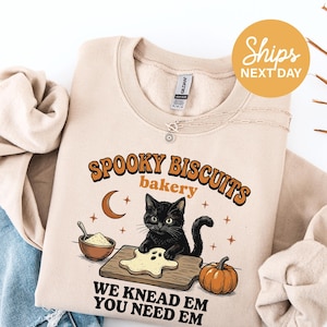 May include: A beige crewneck sweatshirt featuring a graphic of a black cat baking a ghost-shaped cookie on a wooden board. The text reads SPOOKY BISCUITS bakery WE KNEAD EM YOU NEED EM, accompanied by a crescent moon, stars, and a pumpkin.