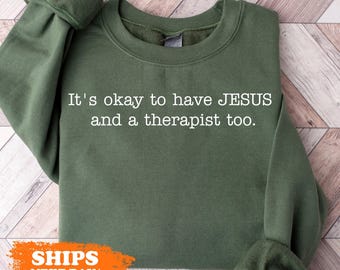 May include: A green sweatshirt with the text "It's okay to have JESUS and a therapist too." printed in white.