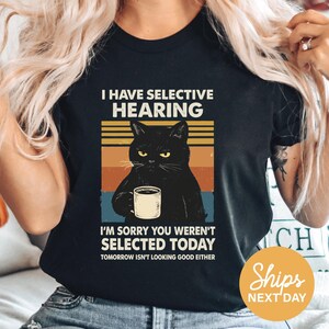 A person wearing a black t-shirt with a graphic design depicting a black cat holding a coffee cup and a humorous caption.